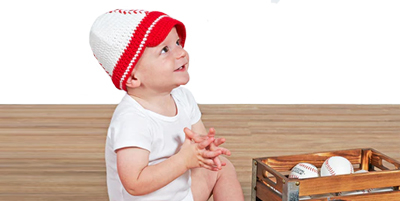 Baby in a baseball knit cap sitting next to a baseball glove and balls