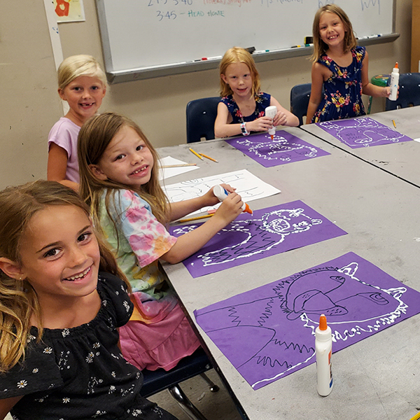 Five girls are sitting around a table working on glue artwork.
