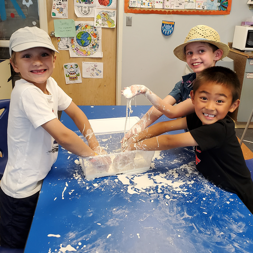 Three boys stand at a table and play in a tub of goop.