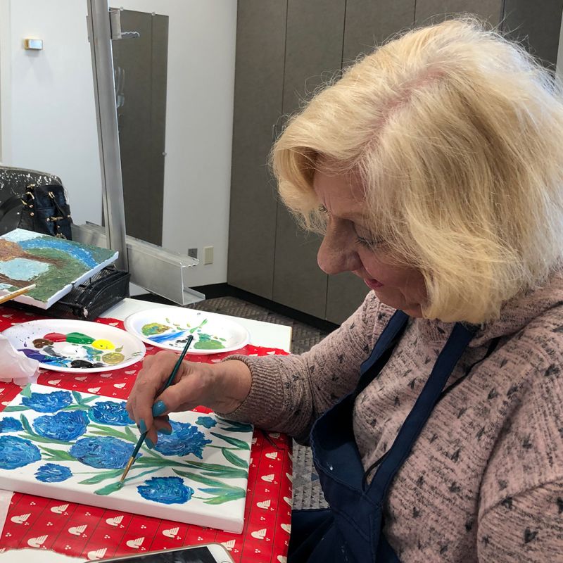 Woman painting on a canvas blue flowers with green stems